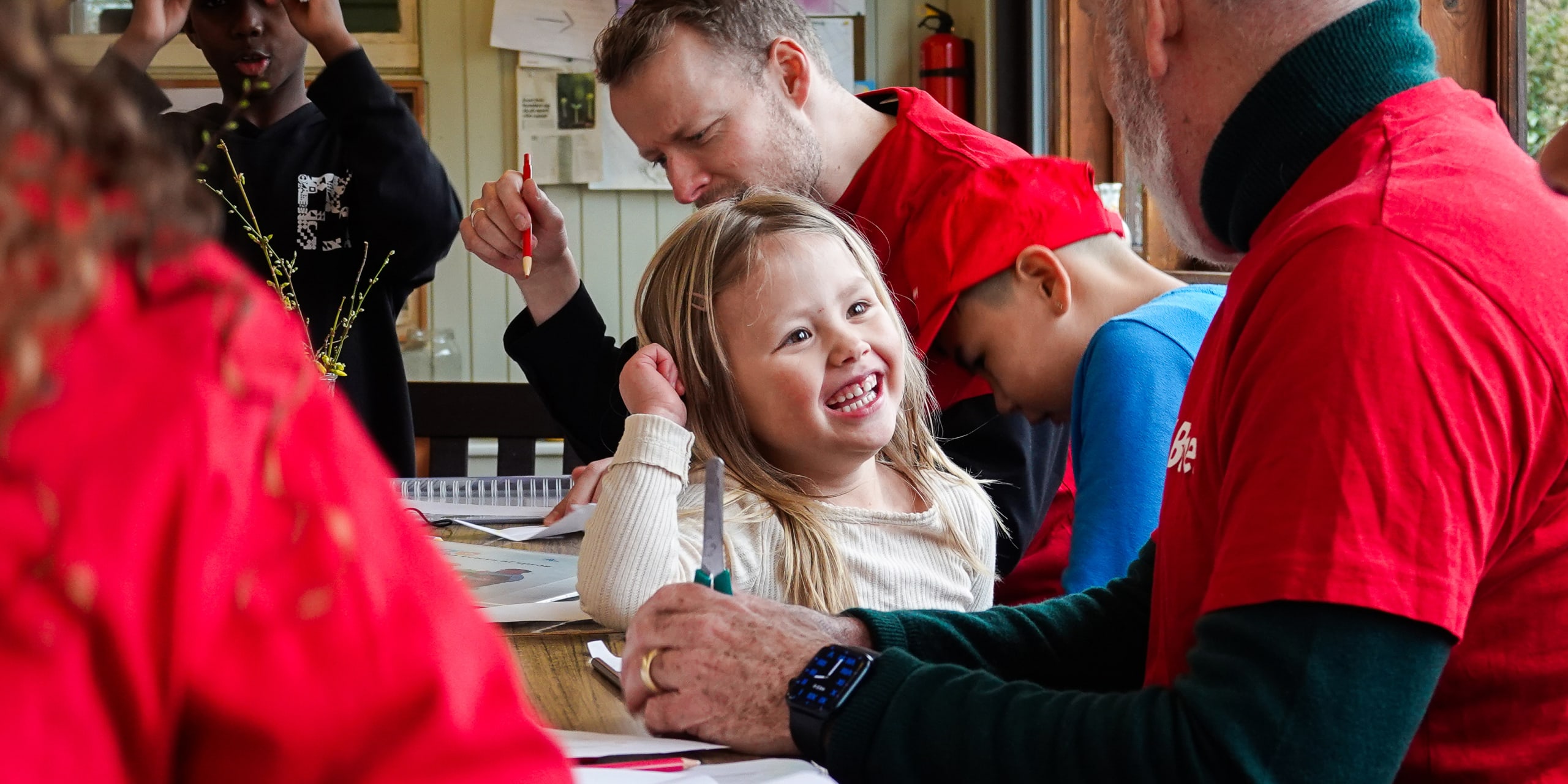 Et lille barn smiler bredt, mens hun deltager i en kreativ aktivitet sammen med andre børn og voksne ved et bord.